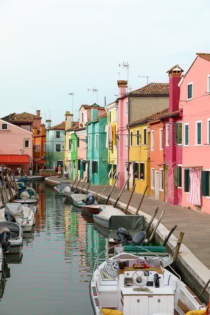 Boats lined up on a canal by a colorful walkway in Burano, Venice