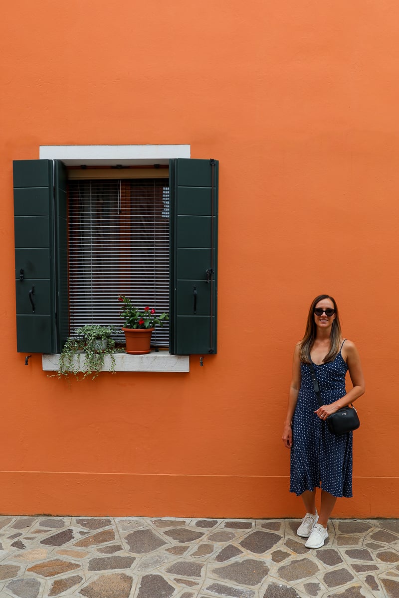 Maddy standing against an orange-painted wall, beside a beautiful window decorated with potted plants, while exploring the streets during the Murano and Burano tours