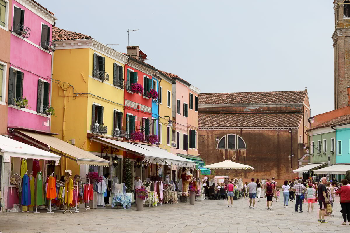 Tourists roaming around a plaza in Burano surrounded by colorful clothing shops