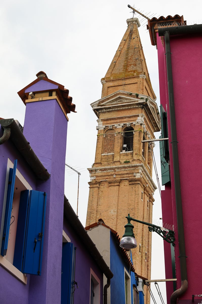 Bell tower standing tall among the colorful houses of Burano. Don't forget to book a day trip to Murano and Burano and enjoy each island's magnificent views.