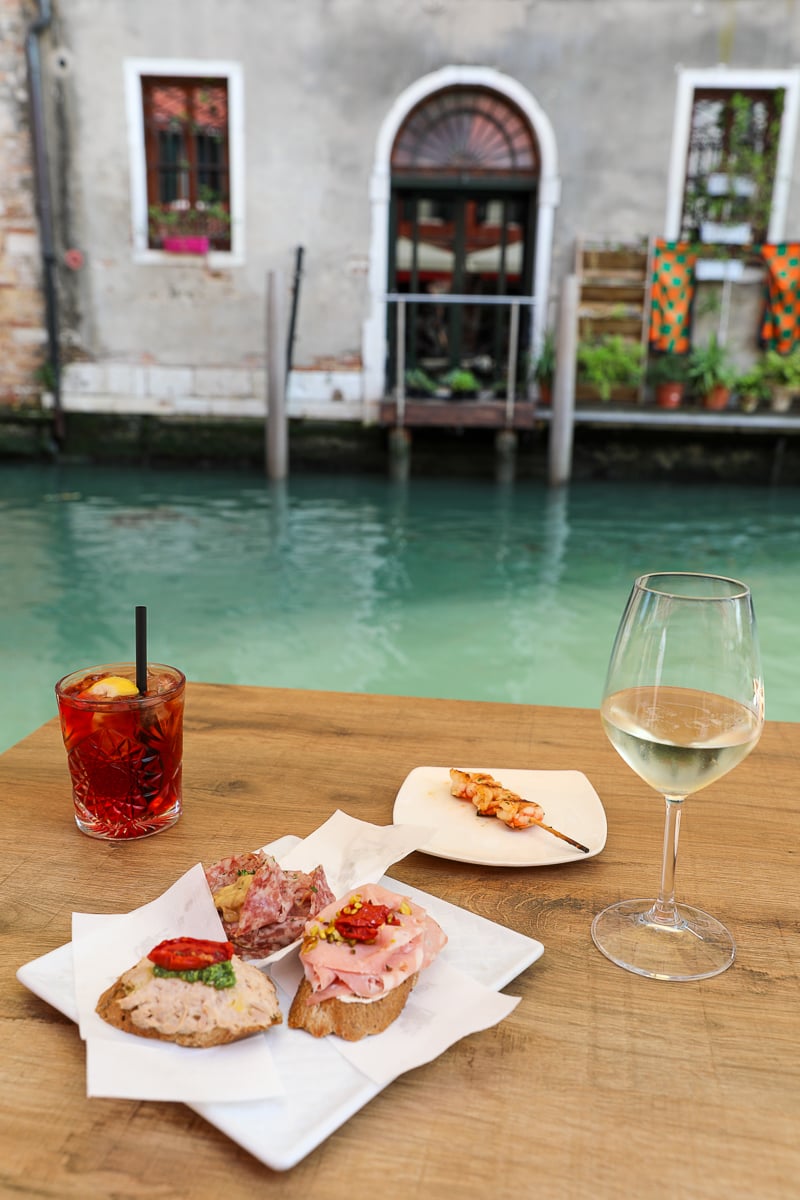 A negroni, a glass of white wine, and some cicchetti snakcs on a table in Venice with a canal in the background