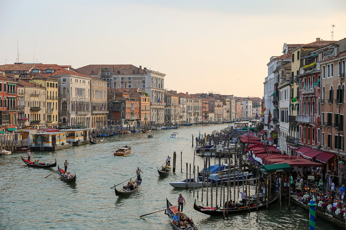 A view overlooking the Grand Canal at Sunset - many tourists are milling about on the side street, while many boats and gondolas flat along the canal