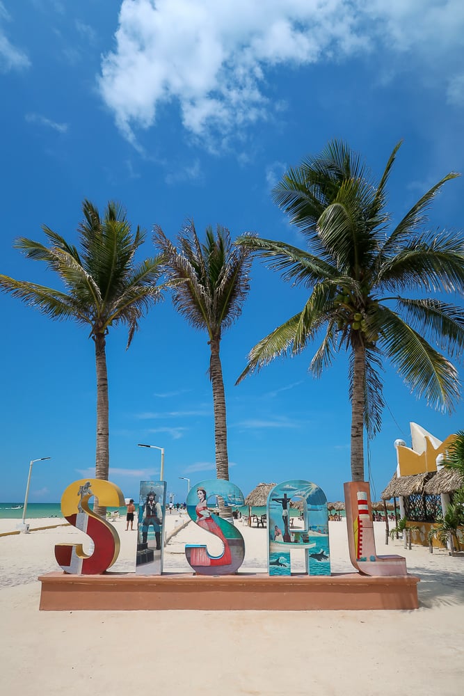 Colorful signage of Sisal by the beach with palm trees behind it. Wondering what to do in Sisal? Roaming the colorful streets and lazing on its white-sand beaches are some of the things to do in Sisal.