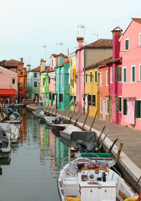 Boats lined up on a canal by a colorful walkway in Burano, Venice