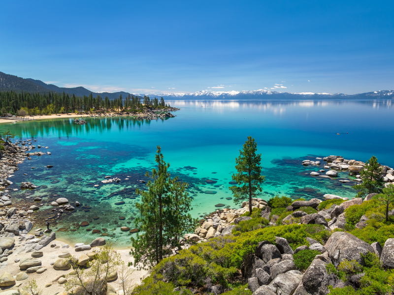 beautiful clear water, rocks, and trees surrounding Lake Tahoe with mountains in the distance. a must-visit destination in california.